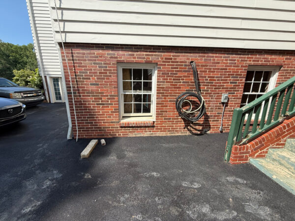 Brick basement wall with a window, a coiled hose on the wall, and a metal staircase with a green railing to the right; parked cars visible on the left.