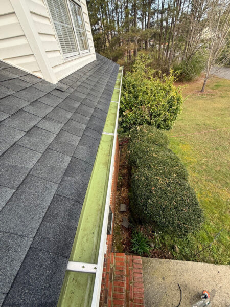 Overhead view of a gray shingled roof with a light-green gutter along the eave, brick wall below and landscaped shrubs nearby.