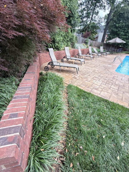 Row of lounge chairs along a brick planter by a swimming pool on a stamped concrete patio, surrounded by greenery and trees.