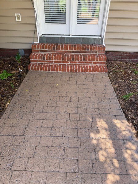Brick steps lead to a white door with glass panels and blinds; a tan-paver walkway extends outward, flanked by garden beds on both sides.