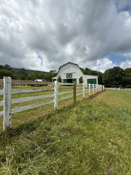 White barn with green doors and trim, set behind a white split-rail fence along a grassy field under a cloudy sky.
