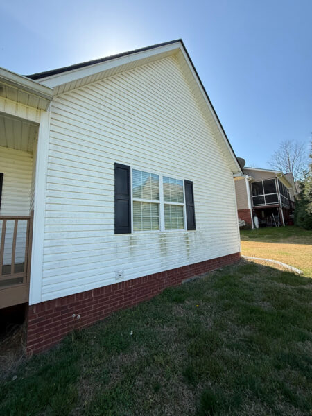 Side view of a white house with vinyl siding, a window with black shutters, and a brick foundation on a sunny day