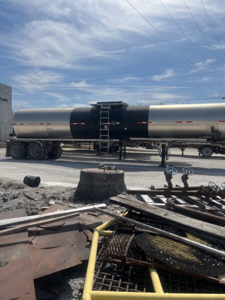 Industrial tanker trailer parked at a loading yard, with a ladder and platform on top and overhead power lines in a blue sky.