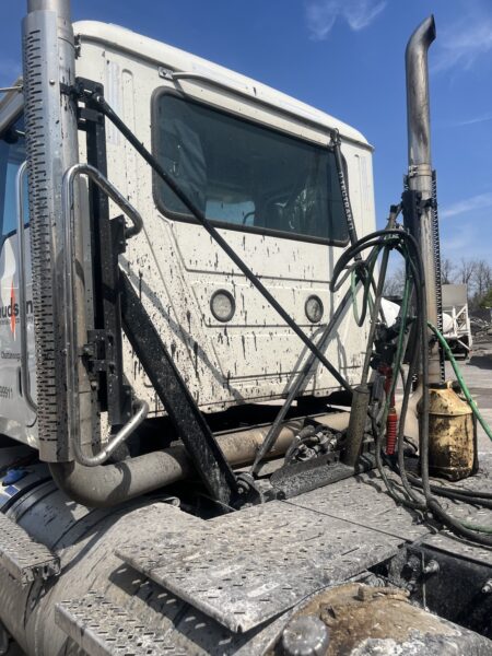 Close-up of a dirty white semi-truck cabin with hoses and metal steps in an industrial area on a sunny day