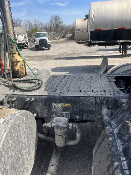 View from inside a dirty tractor-trailer cab showing hoses, a metal platform, and large rear wheels in an industrial yard with other trailers nearby.