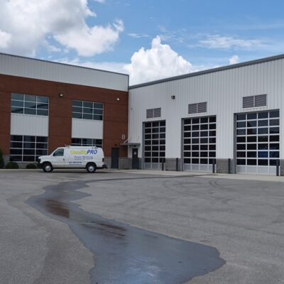 Brick-and-white commercial building with large garage doors and an American flag waving in front; a white van labeled QualityPRO parked nearby on a paved lot.