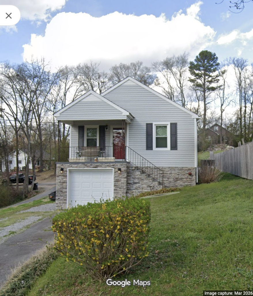 Front view of a gray, single-family house with a red door, stone foundation, and a driveway leading to an attached garage beneath the main level.
