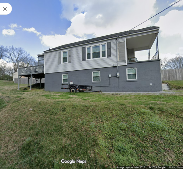 Two-story house with a raised deck on the left and a trailer on the grassy yard in front.