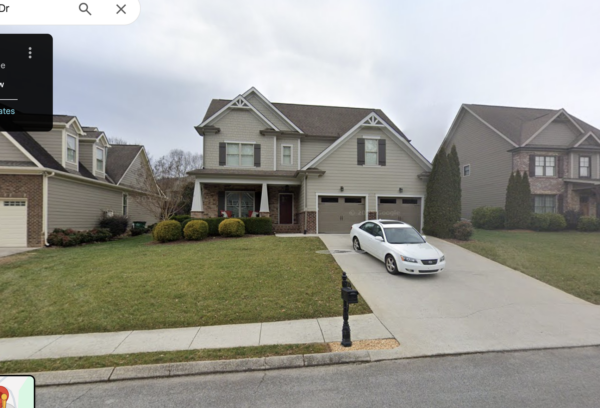 Two-story suburban house with beige siding, a brick porch, and a concrete driveway with a white sedan parked in front of a two-car garage