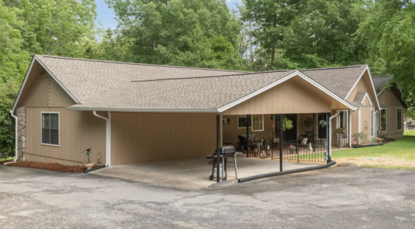 Beige single-story house with a carport, outdoor seating area, and a grill under a covered patio, with green trees in the background.