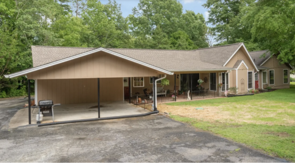 Front view of a beige, ranch-style house with a large covered carport and a front porch, surrounded by green trees.