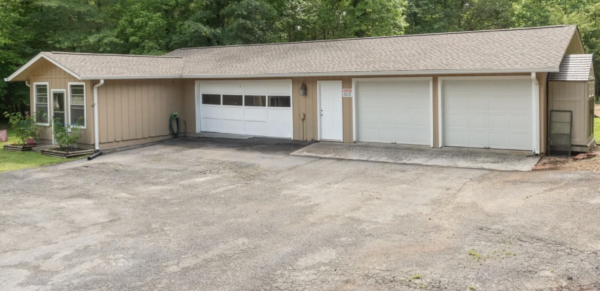 Front view of a beige, single-story building with three white garage doors and a side entry door, surrounded by trees at the back.