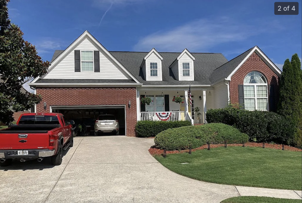 Front view of a two-story brick house with a two-car garage; a red pickup truck is parked in the driveway and the porch is decorated with planters and an American flag.