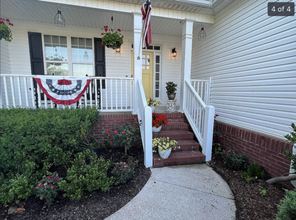 Front porch of a house with white siding, brick steps, and an American flag bunting on the railing beside a yellow front door.