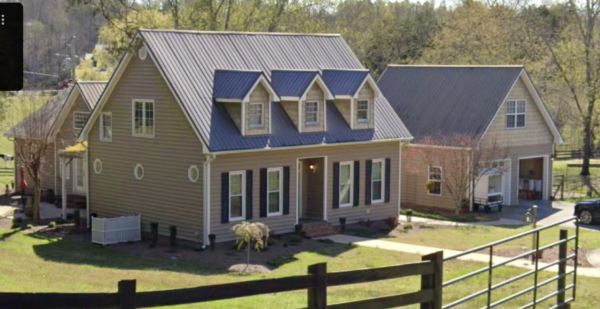 Detached beige house with blue metal roof, dormer windows, and a front porch, set in a fenced yard with trees in the background