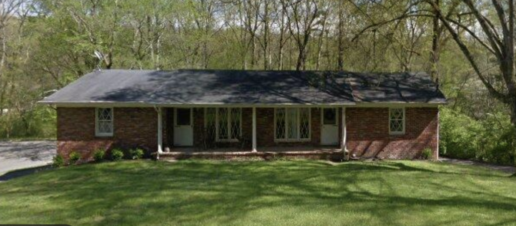 Single-story brick house with a long covered front porch and a grassy, tree-lined yard.
