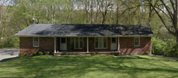 Single-story brick house with a long covered front porch and a grassy, tree-lined yard.