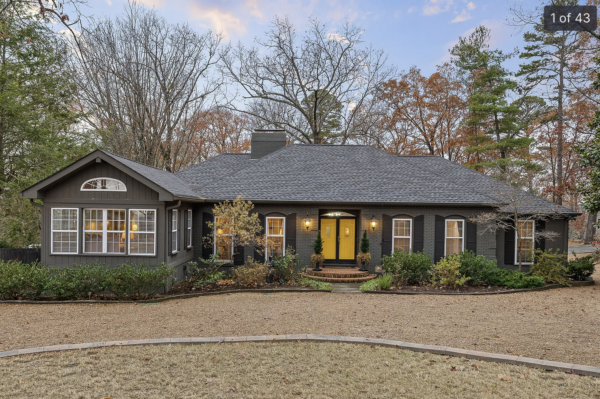 Dark gray brick bungalow with a yellow front door, warm lantern lights, and brick steps framed by landscaped shrubs.