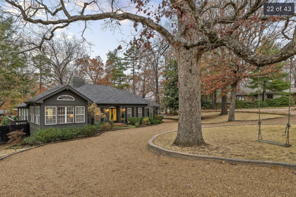 Gray single-story house with a yellow front door sits along a curved gravel driveway, a large tree dominates the yard with autumn leaves on the ground and in the branches.