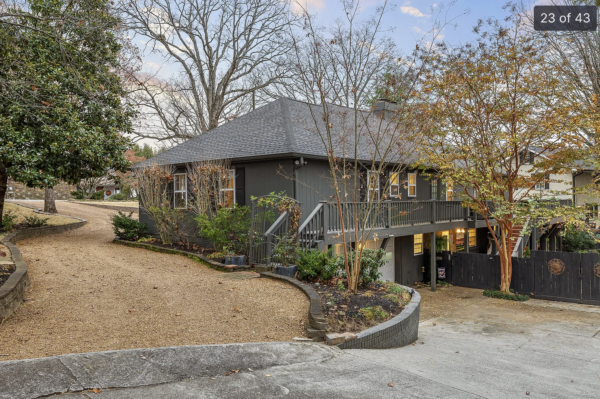 Gray two-story house with a raised porch and stairs, surrounded by trees along a curved gravel driveway, autumn foliage visible on right side.
