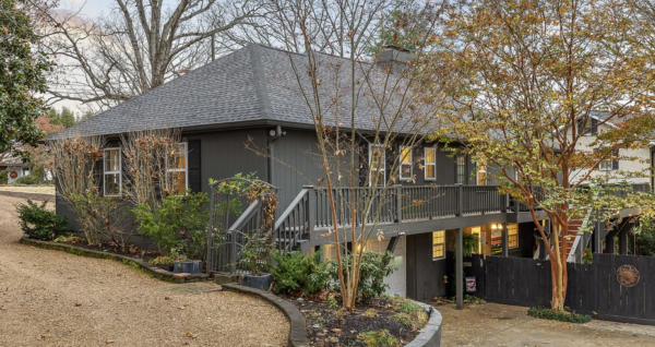 Gray wooden house with a raised deck and steps, surrounded by bare and autumn-colored trees in a residential yard.