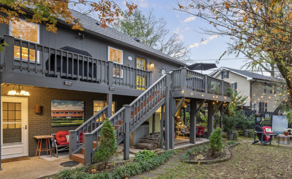Backyard of a two-story gray house with a wooden deck and stairs, string lights, and cozy seating under autumn-colored trees.
