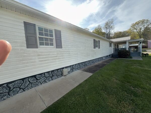 Side view of a white, single-story house with gray shutters along a concrete walkway and a stone foundation, sun glare in the sky.