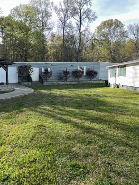 Row of manufactured homes with blue siding, trimmed bushes, and a white mobile home on the right; trees and a wooded backdrop behind.