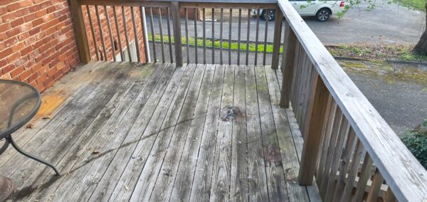 Weathered wooden balcony deck with railing outside a brick building, overlooking a parking lot with a silver car in the distance, leaves scattered on the boards.