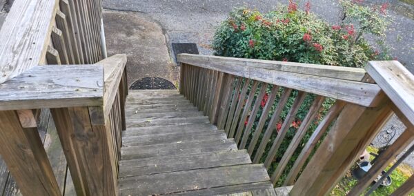 Wooden outdoor staircase with weathered railings, descending toward a small yard and a metal grate at ground level, greenery to the right.