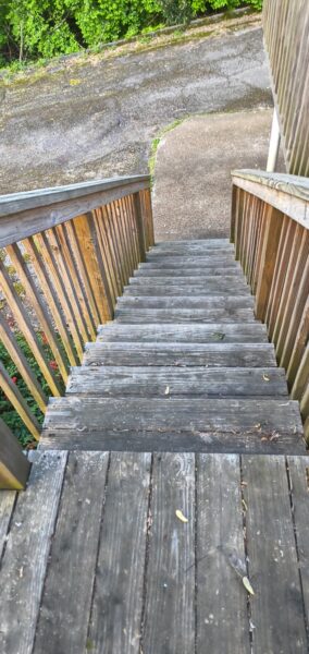 Top-down view of weathered wooden stairs with railings on both sides, leading down to a concrete path.