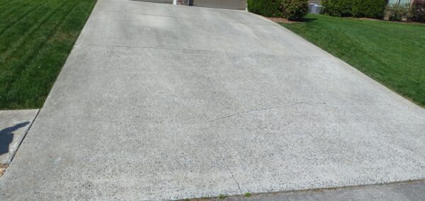Wide concrete driveway with visible cracks leading to a closed garage, flanked by well-manicured green lawns and shrubbery.