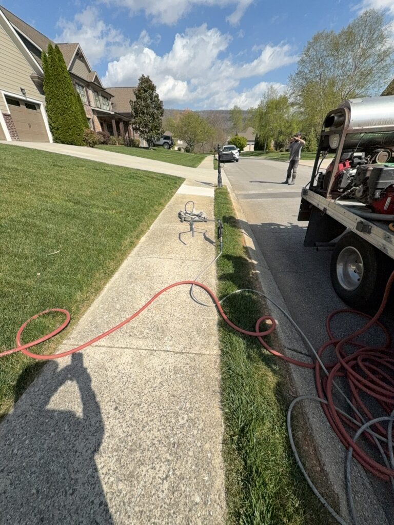 Worker on a suburban street next to a utility truck with tools; red hoses lay across the sidewalk and street.