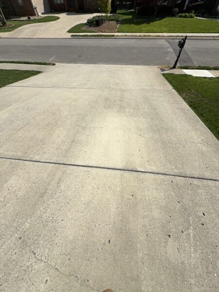 Residential street view with concrete driveway and a leaning mailbox at the curb, green lawns and houses in the background