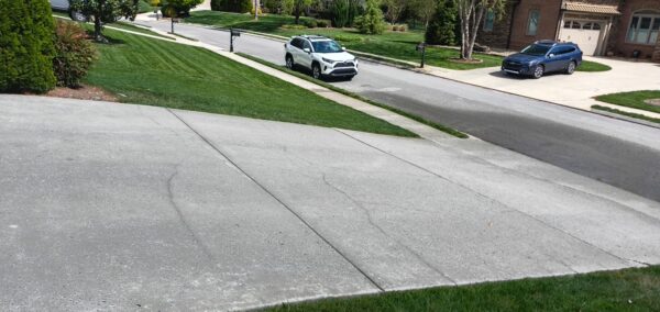 Sloped concrete driveway leads to a suburban street; a white SUV is parked on the curb and a blue SUV sits in a driveway, with green lawns and a brick house in the background.