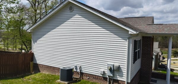 White siding house with a gable roof, visible air conditioner unit and utility meters along the brick foundation in a fenced yard.