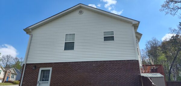 Two-story house with white siding and brick foundation, a wooden deck on the right under a bright blue sky
