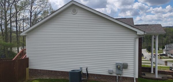 Side view of a white siding house with brick foundation, HVAC unit, and utility boxes in the yard; a wooden fence on the left and a covered porch on the right.