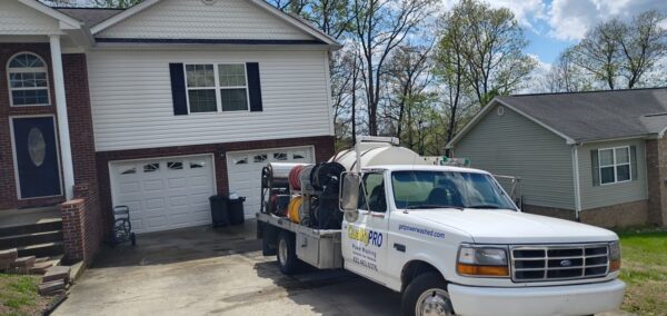 White service truck with hoses and reels loaded, parked in a driveway in front of a two-car garage house in a suburban neighborhood.