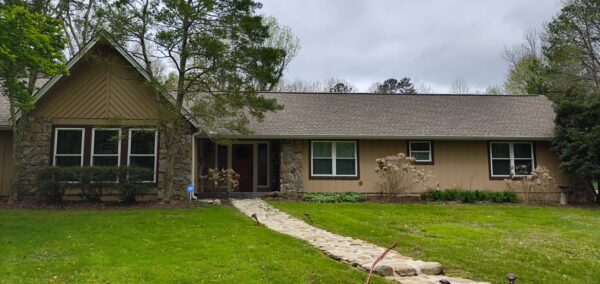 Front view of a ranch-style house with a stone facade, beige siding, and a curved stone walkway leading to the door amid a green lawn and trees under a cloudy sky.