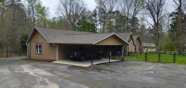 Single-story tan building with a two-car carport housing a dark pickup truck and adjacent yard, trees in the background.