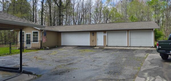 Beige, single-story garage building with three white doors and a side entrance; American flag near a window, wet asphalt driveway, and trees in the background.
