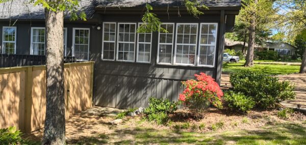 Gray wooden house with white-framed windows, a tall tree in front, and a bright red azalea bush by the entry area behind a natural wooden fence.