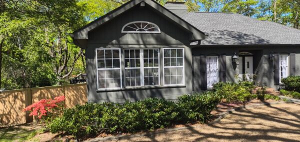 A dark-gray house with large front bay windows and a gravel walkway bordered by green shrubs and a pink flowering bush in front.