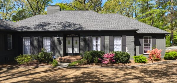 A single-story gray brick house with a double front door, brick steps, and black shutters, surrounded by green shrubs and flowering azaleas on a sunny day.