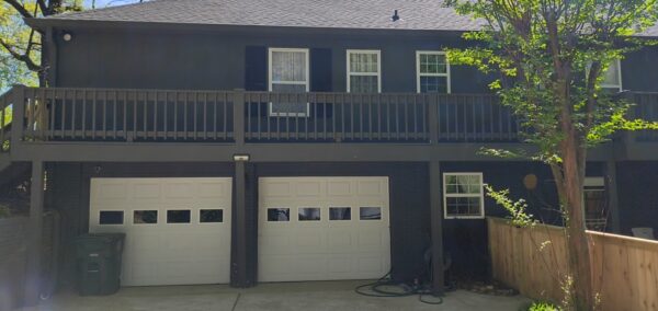 Two-story blue townhouse with a wooden balcony, two white garage doors, and a tree on the right.