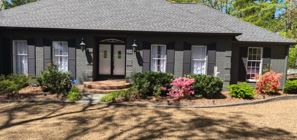 Front view of a single-story gray brick house with black shutters, double doors, and brick steps, bordered by flowering shrubs.