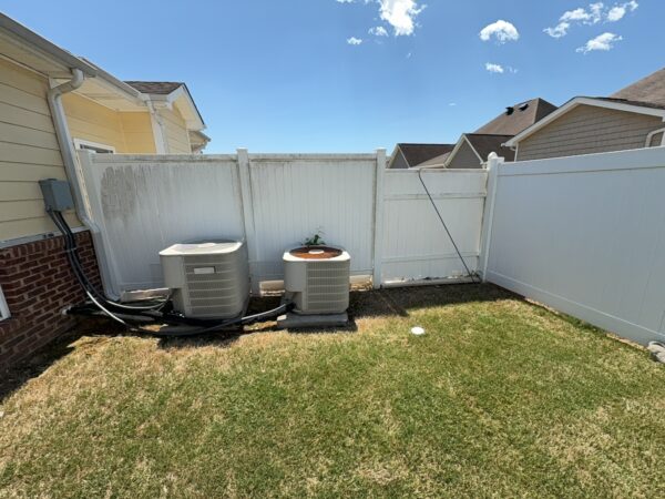 Backyard with two outdoor AC units on concrete pads beside a white vinyl fence and yellow house wall under a blue sky.