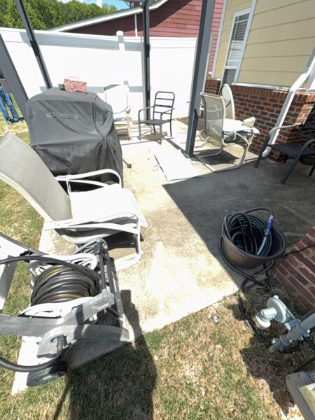 Patio area with a covered grill, several chairs, and tangled hoses and cables on a sunny day beside a brick wall and white fence.