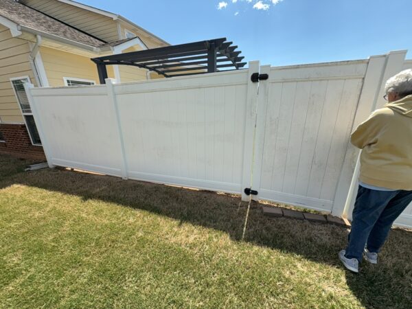 Man in a yellow jacket stands near a white vinyl gate with a black latch in a sunny yard.